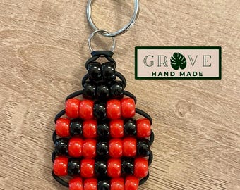May include: Handmade ladybug keychain. The keychain features a ladybug design made of red and black beads. The keychain is attached to a silver keyring. The text "GROVE HAND MADE" is in a green rectangle.