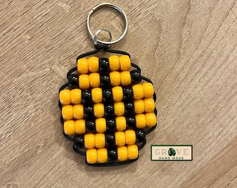 May include: A handmade keychain featuring a square design made of yellow and black beads. The beads are strung together with black string, and attached to a silver key ring. The keychain has the text "GROVE HAND MADE" on a green and white label.
