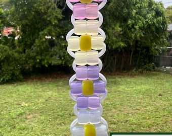 May include: A handmade, vertical beaded ornament with alternating sections of lavender, white, and yellow beads. The ornament is suspended by a clear loop and features the text "GROVE HAND MADE" in a green and white logo.