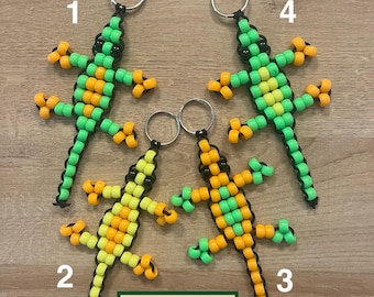 May include: Four beaded lizard keychains in green, yellow, and orange. Each lizard has a black beaded outline and a silver keyring. The keychains are arranged on a wooden surface. The text "GROVE HAND MADE" is on a green rectangle.