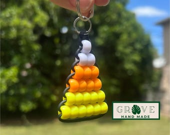 May include: A handmade keychain featuring a candy corn design made of yellow, orange, white, and black beads. The keychain is attached to a silver keyring. The brand name "GROVE HAND MADE" is displayed on a green and white label.