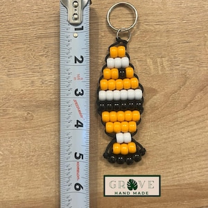 May include: A handmade beaded keychain in a fish shape, featuring orange, white, and black beads. The keychain is approximately 3 inches long. The design includes a metal keyring and the text "GROVE HAND MADE" on a green and white label.