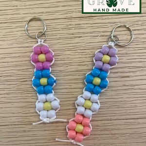 May include: Two handmade beaded flower keyrings. Each keyring features a flower design with colourful beads in shades of pink, blue, white, and yellow. The keyrings are attached to silver key rings. The text "GROVE HAND MADE" is in a green rectangle.