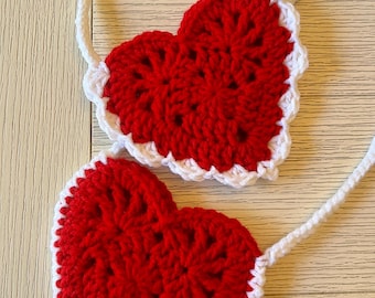 May include: Two handmade crochet heart-shaped items. Each heart is red with a white scalloped border and a white cord. The items are on a light wood surface. The text "GROVE HAND MADE" is in a green and white rectangle.