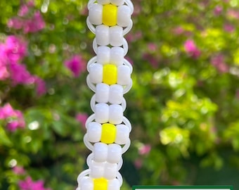 May include: A handmade daisy chain keychain with white and yellow beads. The keychain is attached to a silver ring. The background is a blurred view of green foliage and pink flowers. The keychain has the text "GROVE HAND MADE" on a green rectangle.