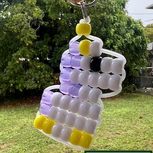 May include: A handmade keychain with a design of yellow, white, black, and purple beads. The keychain is attached to a silver metal ring. The bottom of the image shows a green and white sign that reads "GROVE HAND MADE".