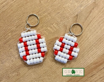 May include: Two handmade baseball keychains. Each keychain features a baseball design made of white and red beads. The keychains are attached to silver key rings. The keychains are on a wooden surface. The text "GROVE HAND MADE" is on a green and white label.