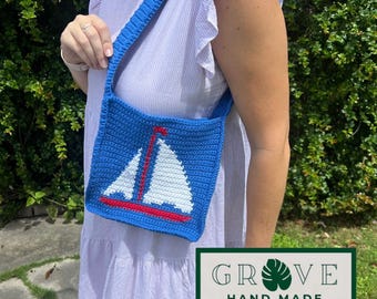 May include: A blue crochet shoulder bag featuring a white sailboat design with red accents. The bag has a matching blue strap and is worn over the shoulder. The bag is handmade and the text "GROVE HAND MADE" is visible.