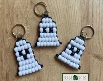 May include: Three handmade ghost keychains. Each keychain features a ghost design made of white and black beads, with a gold-colored keyring attached. The keychains are arranged on a wooden surface, with a green and white logo in the bottom right corner.
