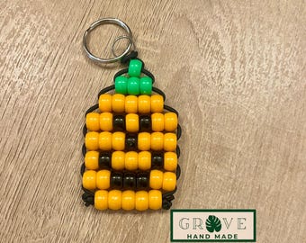 May include: A handmade beaded pineapple keychain. The pineapple is made of yellow and black beads, with a green beaded top. The keychain has a silver ring and the text "GROVE HAND MADE" on a green and white label.