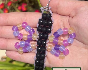 May include: A handmade butterfly keychain made of colorful beads. The butterfly has black beads for the body and wings in shades of purple, pink, and yellow. The keychain includes a silver-colored metal ring and the text "GROVE HAND MADE".