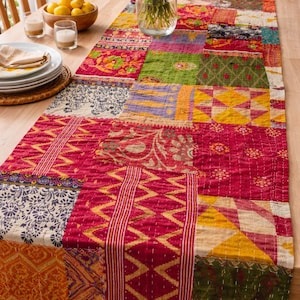 Könnte beinhalten: Ein farbenfroher Patchwork-Tischläufer mit verschiedenen Mustern und Farben, darunter Rot, Gelb, Grün und Blau. Der Läufer liegt auf einem Holztisch, auf dem eine Vase mit Blumen steht.