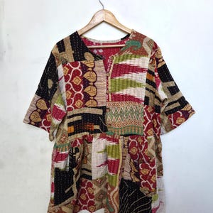 May include: A patchwork tunic top with short sleeves, hanging on a wooden hanger. The top features a variety of fabric patterns in red, black, beige, and green. It has a V-neckline and button closure.