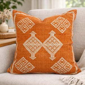Könnte beinhalten: Ein quadratisches Dekokissen in leuchtendem Brandorange mit einer strukturierten Webart. Das Kissen ist mit einem cremefarbenen, gestickten geometrischen Muster verziert, das aus vier Eckquadraten und zwei zentralen Rautenformen besteht.