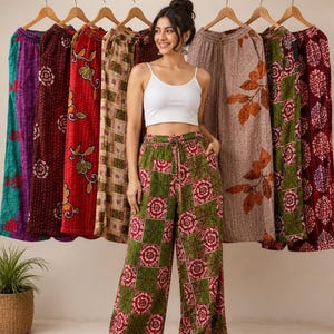 May include: A collection of wide-leg trousers featuring various patterns and colors, including red, green, brown, and purple, displayed on wooden hangers. The trousers have an elastic waistband with a drawstring and are made of quilted fabric.