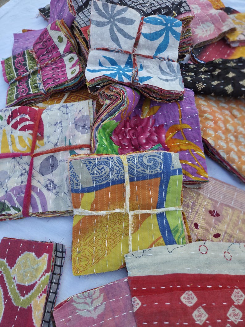 May include: A collection of colorful, patterned fabric bundles tied with string. The fabrics feature floral and geometric designs in shades of pink, purple, yellow, blue, and orange. The bundles are stacked together, showcasing the variety of patterns.