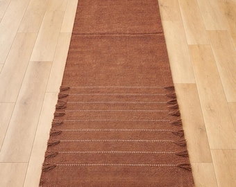 May include: A rectangular, brown woven rug with a textured surface. The rug features horizontal white stitching and fringe detailing on the ends. The rug is placed on a light-colored wooden floor.