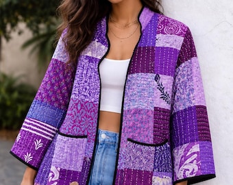 May include: A quilted jacket featuring a patchwork design in various shades of purple, including lavender, violet, and deep plum. The garment has three-quarter length sleeves, a black piped trim along the edges, and two front patch pockets.