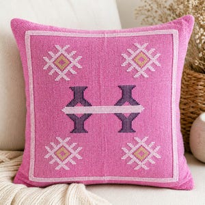 May include: A square throw cushion in a vibrant pink hue featuring a white embroidered border. The centre displays a dark purple geometric motif, while each corner is accented with a white and gold cross-stitch pattern on the textured fabric.