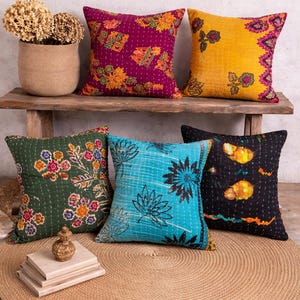 Könnte beinhalten: Fünf dekorative Zierkissen mit floralen Mustern in verschiedenen Farben, darunter Lila, Gelb, Grün und Blau. Die Kissen sind auf einer Holzbank und einem gewebten Jute-Teppich platziert. Eine Vase mit Trockenblumen ist ebenfalls zu sehen.