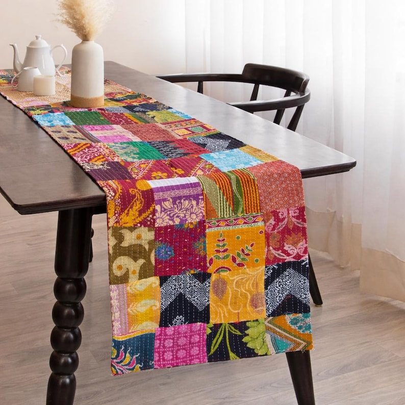 Puede incluir: Un camino de mesa de patchwork colorido con una variedad de cuadrados de tela estampada en tonos rojos, amarillos, azules y verdes. El camino de mesa se coloca sobre una mesa de madera oscura, agregando un toque vibrante al comedor. Mide aproximadamente 183 cm de largo.