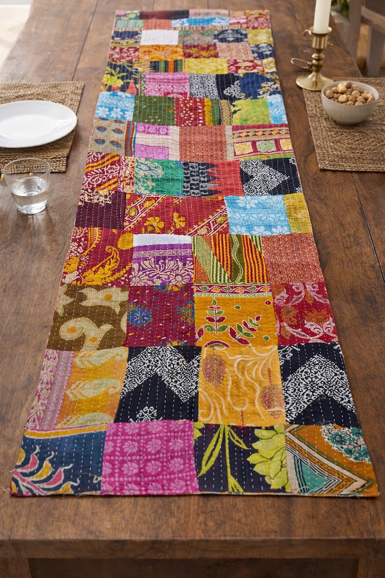 Puede incluir: Un camino de mesa de patchwork colorido con una variedad de cuadrados de tela estampada. El camino de mesa presenta una mezcla de colores vibrantes, incluyendo rojo, amarillo, azul y verde. El camino de mesa est&aacute; colocado sobre una mesa de madera.