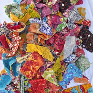 May include: A pile of colorful fabric scraps with various floral and geometric patterns. The scraps are in shades of red, orange, yellow, green, blue, and purple. The fabric appears to be hand-stitched, with visible thread lines.