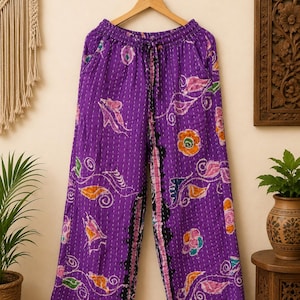 May include: Wide-leg pants in a vibrant purple hue featuring a white kantha stitch pattern and a colorful floral print. The pants have an elastic waistband with a drawstring tie and a black vertical decorative trim running down the inner legs.