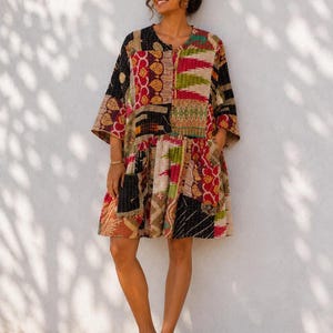 Könnte beinhalten: Ein Patchwork-Kleid mit rundem Ausschnitt und Dreiviertelärmeln. Das Kleid hat eine Vielzahl farbenfroher Muster in Rot, Schwarz, Grün und Beige. Das Kleid hat einen gerafften Rock und Taschen. Die Person trägt braune Sandalen.