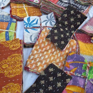 May include: A collection of colorful, patterned fabric squares, likely textiles or quilts. The fabrics display a variety of designs, including floral, geometric, and paisley patterns, in shades of orange, brown, blue, and purple. The pieces are stacked and bundled.