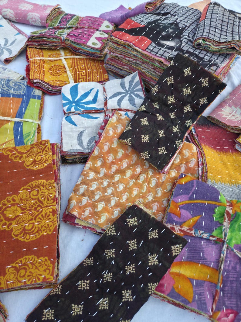 May include: A collection of colorful, patterned fabric squares, likely textiles or quilts. The fabrics display a variety of designs, including floral, geometric, and paisley patterns, in shades of orange, brown, blue, and purple. The pieces are stacked and bundled.