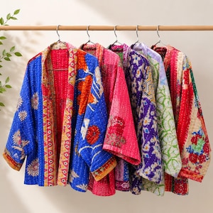 Könnte beinhalten: Fünf gesteppte Jacken hängen an einer Holzstange, jede mit unterschiedlichen lebendigen Mustern und Farben wie Blau, Rot, Pink, Lila und Grün. Die Kleidungsstücke haben eine offene Vorderseite, lange Ärmel und sichtbare Kantha-Steppnähte.