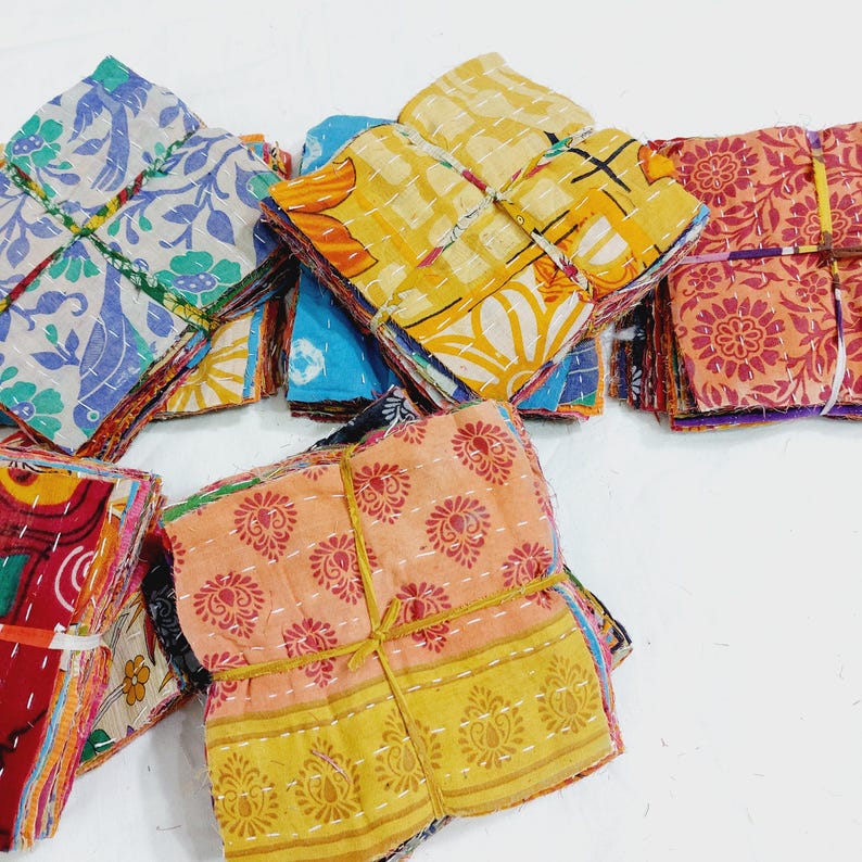 May include: Stacks of colorful, patterned fabric squares tied with twine. The fabrics feature floral and geometric designs in shades of blue, yellow, orange, and red. The squares are likely part of a textile craft project.