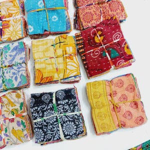 May include: A collection of colorful, patterned fabric squares, bundled and tied with twine. The fabrics feature floral and geometric designs in shades of blue, yellow, red, and orange. The squares are stacked and ready for use.