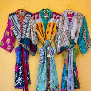 Könnte beinhalten: Drei farbenfrohe Patchwork-Kimono hängen an gelben Kleiderbügeln vor einem gelben Hintergrund. Die Kimonos weisen eine Vielzahl von Mustern und Farben auf, darunter Blumen-, geometrische und abstrakte Designs. Der Text "Patchwork Kimonos" steht oben.