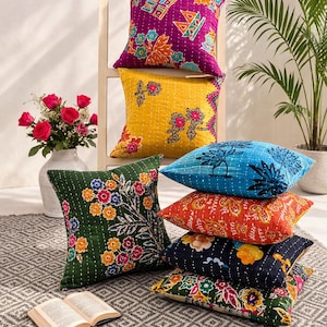 Könnte beinhalten: Eine Ansammlung farbenfroher Zierkissen mit floralen und geometrischen Mustern. Die Kissen sind in Lila, Gelb, Blau, Orange und Grün gehalten. Sie liegen auf einem gemusterten Teppich, mit einem Buch und einer Vase mit Rosen in der Nähe.