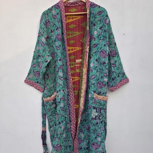 Può includere: Accappatoio stile kimono turchese con motivo floreale, dettagli viola e interno rosso a contrasto. L'accappatoio ha tasche e una cintura abbinata. Il capo è esposto su una gruccia di legno su sfondo bianco.