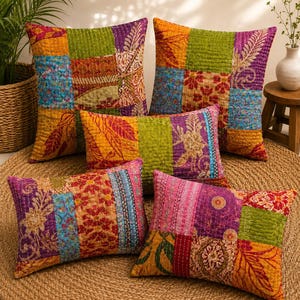Könnte beinhalten: Eine Sammlung von fünf quadratischen und rechteckigen Dekokissen mit einem Patchwork-Design aus leuchtenden Stoffen in Grün-, Orange-, Lila-, Blau- und Rosatönen. Jedes Kissen ist mit sichtbaren weißen Kantha-Nähten über die gesamte Fläche versehen.