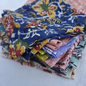 Könnte beinhalten: Ein Stapel bunter Stoffservietten mit floralen Mustern. Die Servietten weisen eine Vielzahl von Farben auf, darunter Blau, Rosa, Gelb und Grün. Die floralen Designs umfassen große, stilisierte Blumen und Blätter.