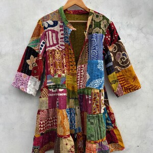 May include: A patchwork jacket with a variety of colorful fabric squares. The jacket has a collar and three-quarter sleeves. The garment is hanging on a wooden hanger against a neutral background.