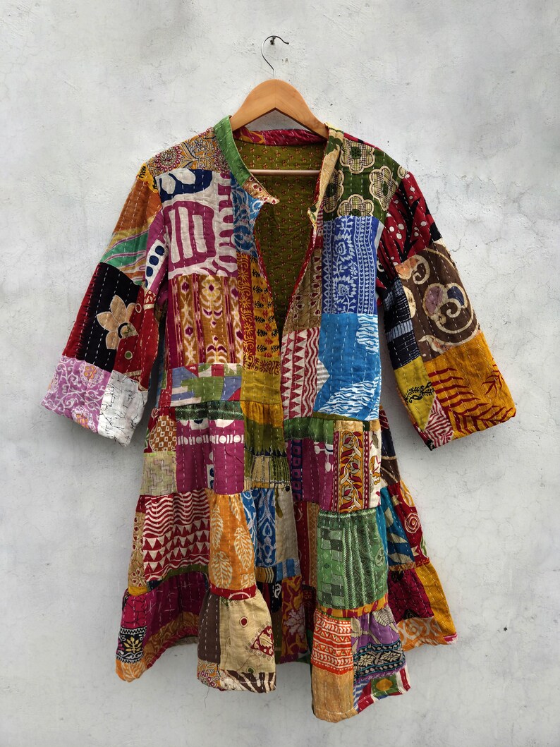 May include: A patchwork jacket with a variety of colorful fabric squares. The jacket has a collar and three-quarter sleeves. The garment is hanging on a wooden hanger against a neutral background.
