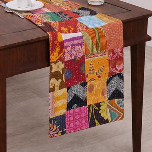 Puede incluir: Un colorido camino de mesa patchwork con una variedad de cuadrados de tela estampada en tonos rojos, naranjas, amarillos y azules. El camino de mesa est&aacute; colocado sobre una mesa de madera oscura. Un plato blanco, cubiertos y una vela tambi&eacute;n est&aacute;n presentes.