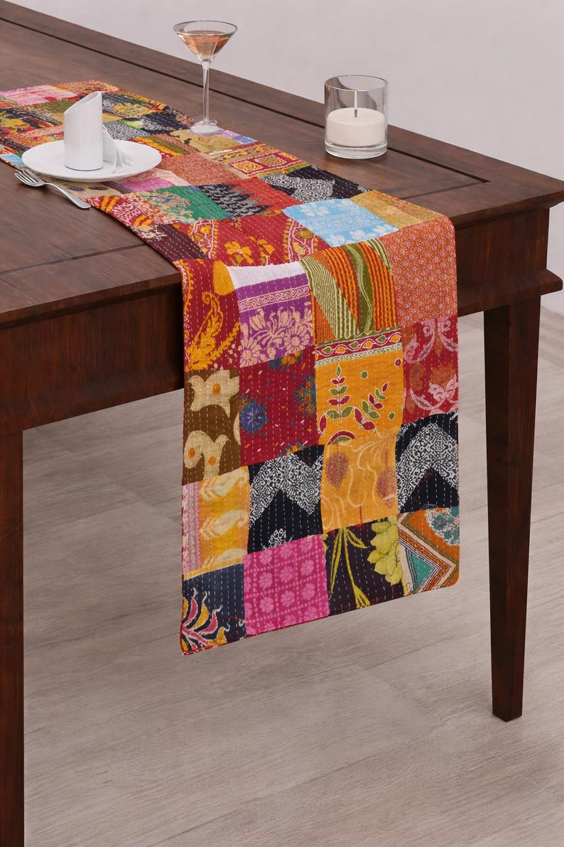 Puede incluir: Un colorido camino de mesa patchwork con una variedad de cuadrados de tela estampada en tonos rojos, naranjas, amarillos y azules. El camino de mesa est&aacute; colocado sobre una mesa de madera oscura. Un plato blanco, cubiertos y una vela tambi&eacute;n est&aacute;n presentes.