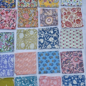 May include: A collection of folded fabric squares with various floral and botanical patterns. The squares feature a range of colors, including blue, pink, yellow, and green, with detailed designs. The fabric appears to be cotton or a similar material.