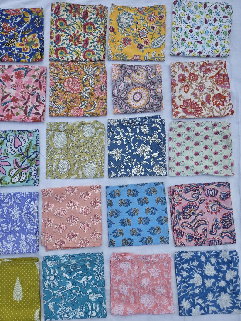 May include: A collection of folded fabric squares with various floral and botanical patterns. The squares feature a range of colors, including blue, pink, yellow, and green, with detailed designs. The fabric appears to be cotton or a similar material.