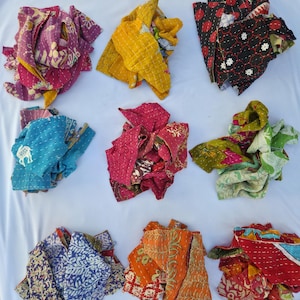 May include: Nine colorful, patterned fabric scraps in various colors, including purple, yellow, red, black, blue, pink, orange, and green. The fabrics appear to be quilted or stitched with decorative patterns. The scraps are arranged on a white surface.