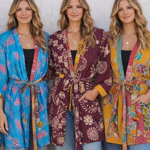 Könnte beinhalten: Drei Frauen tragen farbenfrohe Kimono-Jacken mit Mustern. Die Jacken sind in Blau, Burgunderrot und Gelb gehalten und weisen florale und geometrische Designs auf. Jede Jacken hat einen passenden Gürtel und wird mit Jeans kombiniert.
