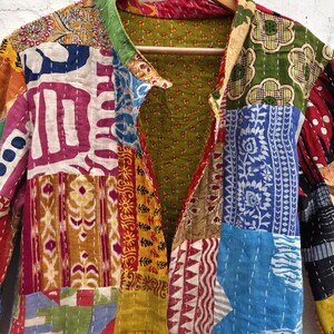 May include: A patchwork jacket with a variety of colorful fabric patterns. The jacket is hanging on a wooden hanger. The design includes floral, geometric, and abstract shapes in shades of red, blue, green, and yellow.