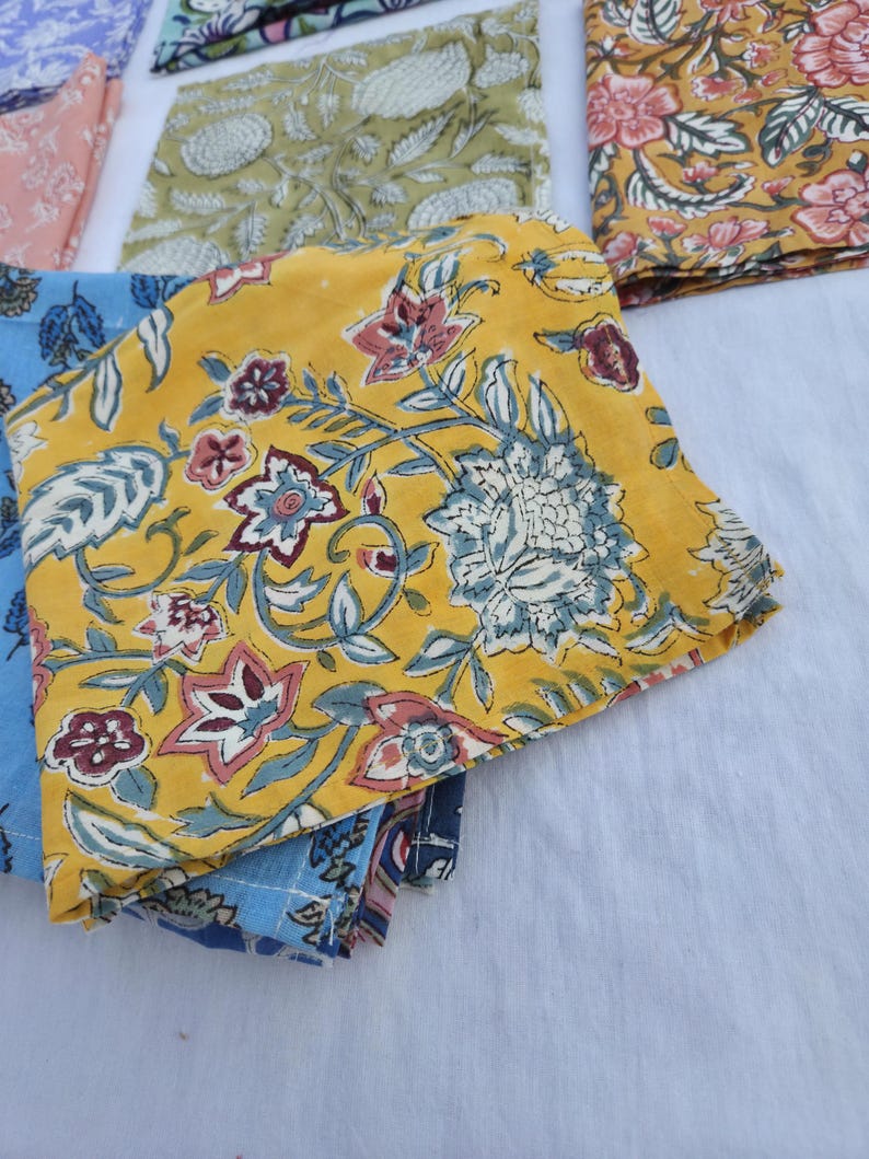 May include: Folded fabric napkins with floral patterns. The napkins feature a variety of colors, including yellow, blue, and pink. The floral designs include leaves and flowers. The napkins are made of a soft material.