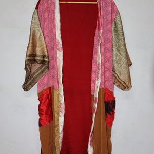 May include: A long, open-front robe featuring a patchwork design with various fabric panels in shades of deep red, pink, brown, and gold. The garment includes wide sleeves, a solid red back panel, and a cream-colored trim with a subtle floral pattern.